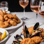 Plates of stuffed mussels and fried shrimp on a table with glasses of rosé wine for the article titled, "The Best Wine to Drink with Seafood."