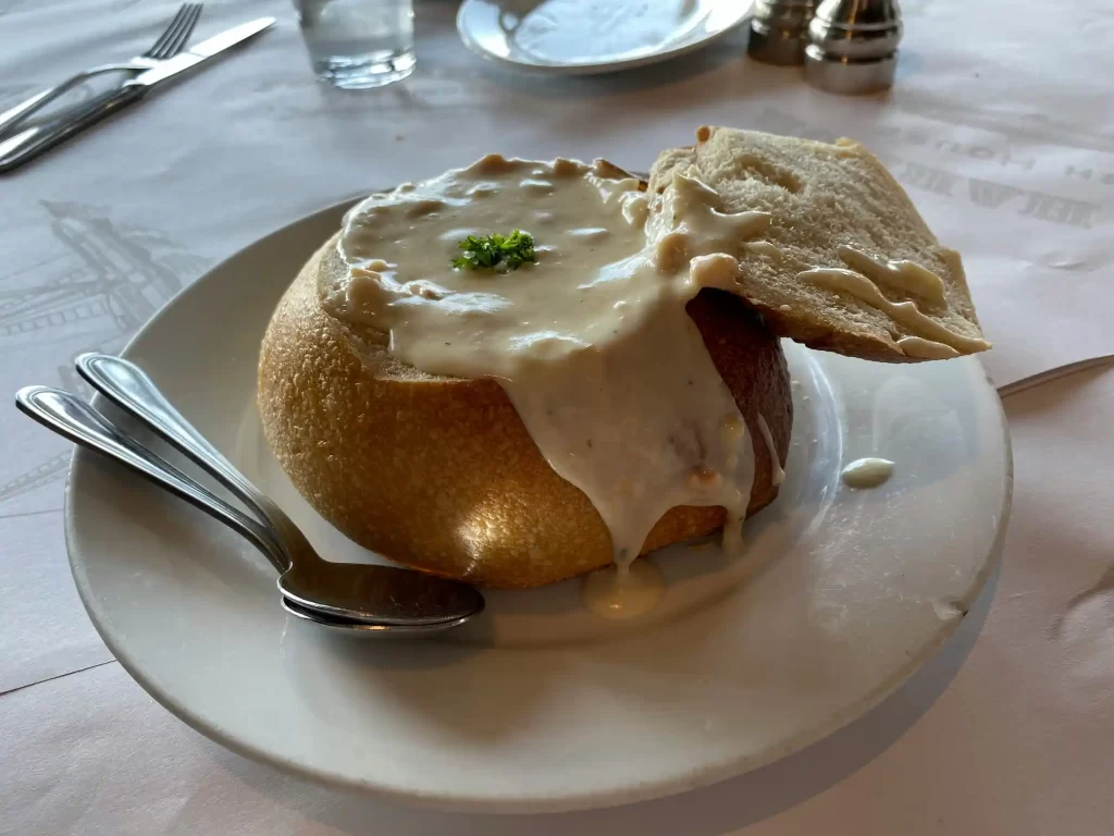 A soup bread bowl containing clam chowder on a white ceramic plate with two spoons.
